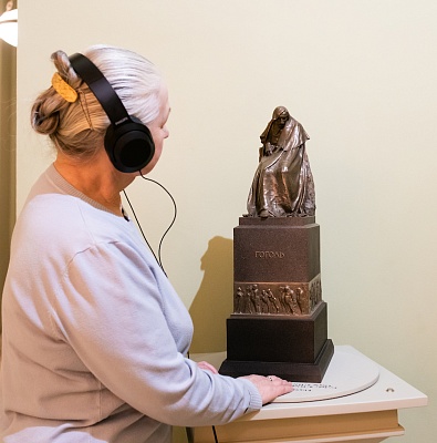 Посетительница слушает аудиогид к тактильному макету памятника, надев наушники, стоя у модели и читая этикетку