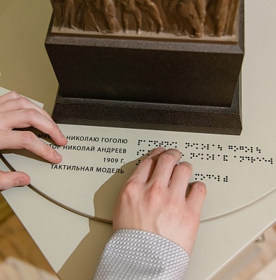 Руки касаются этикетки, набранной шрифтом Брайля на постаменте тактильного макета памятника Николаю Гоголю